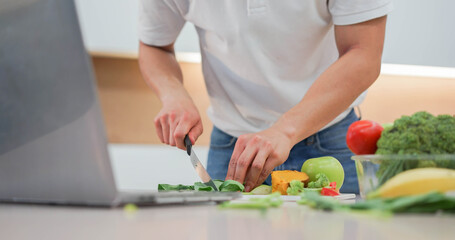 close up man hand using knife to cut vegetables on board and preparing food while open laptop to...