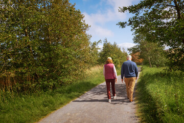 Old couple walking  on a foot path on a warm sunny day. Cloudy sky, Green trees along walking way.