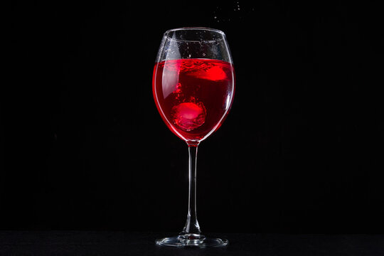 A Glass Of Red Vine On Black Background. Wine Glass On Dark Table. Falling Ice Cubes And Splashes