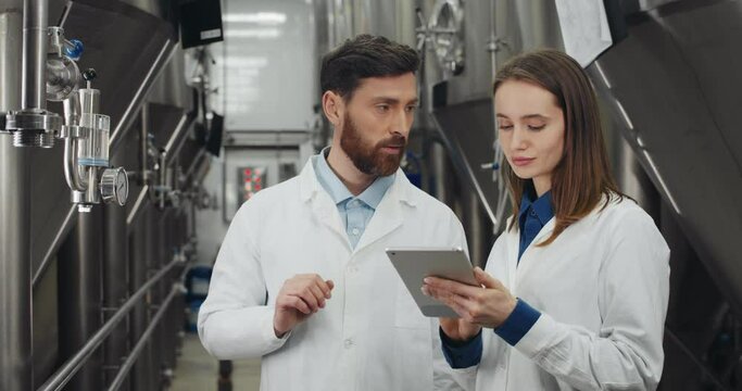 Process engeneer and his assistant looking at equipment gauge and talking. Man and woman in white lab coats entering data and using tablet while standing at rows of brewing vats