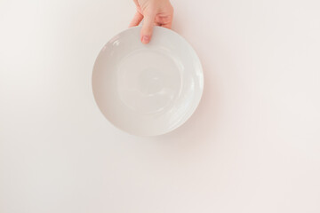  Female hand holds a white round plate on a white background as a mockup with copy space. Ceramic flat saucer