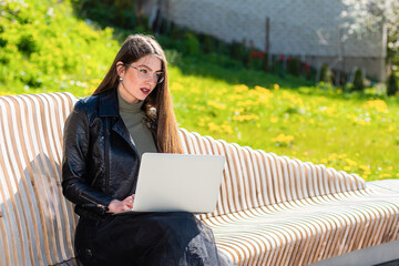 young elegant woman in eyeglasses sitting on bench in park and working on laptop on a sunny and windy day