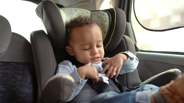 Little mixed-race boy sitting in child car seat and playing with belts. Arfican American toddler ready for travelling in car. Safety and transportation concept