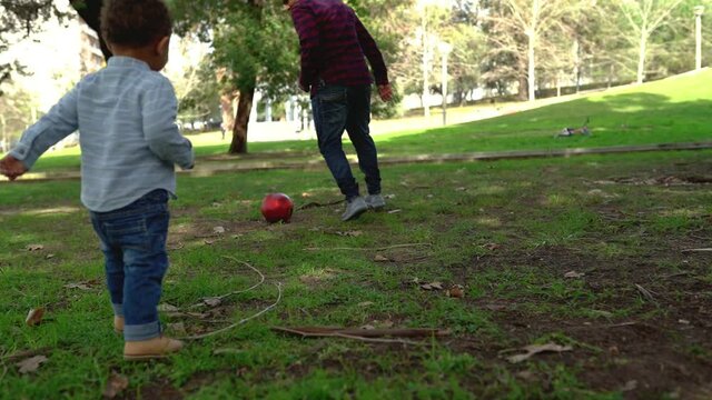 Little African American boy playing football with dad in park. Bald father running on lawn with little son and kicking ball. Family, summer and game concept