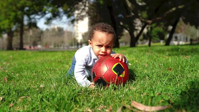 Little African American boy playing with ball in park. Cute toddler crawling on grass, trying to rise ball up from ground and biting it. Childhood, summer and game concept