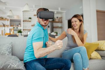 Fototapeta premium Smiling young man using VR headset at home on couch. Man and his wife enjoying virtual reality at her apartment. 