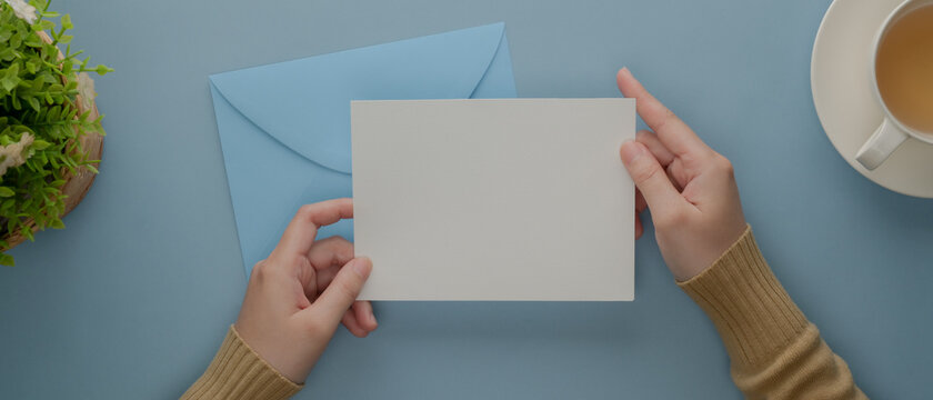 Female hands holding mock-up greeting card above blue envelope on light blue desk