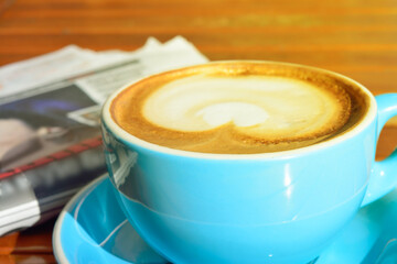 Heart-shaped pattern in a cup of cappuccino with newspaper and wooden table in background.