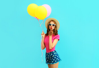 Summer colorful image of cute young woman with yellow pink balloons blowing red lips sending sweet air kiss wearing a shorts and straw hat on blue wall background