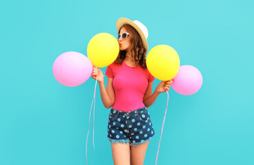 Fototapeta na wymiar Summer colorful image of cute young woman kissing yellow pink balloons wearing a shorts and straw hat on blue wall background