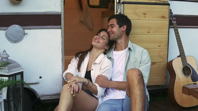 Lovely portrait of a young couple. They are sitting on the stairs of their modern trailer, embracing and smiling. Dreaming and thoughtful mood