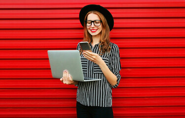 Modern young woman holding laptop looking at phone on red wall background