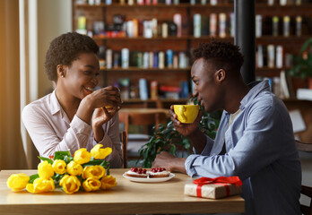 Romantic holiday celebration. African American couple with gift box and flowers drinking coffee in city cafe