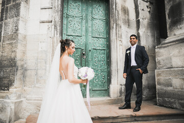 Wedding photo shooting. Bride and bridegroom walking in the city. Married couple embracing and looking at each other. Holding bouquet