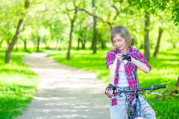 Senior woman spraying insect repellent or sunscreen lotion on her skin while ride a bike at summer park. Empty space for text