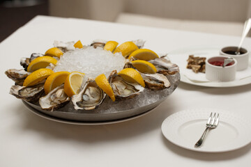 side view close up food shot of fresh raw shucked open oysters lying between lemon slices on a round cold ice tray next to Mignonette shallot vinegar and Ponzu sauces on a white table