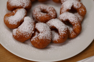 a pile of delicious sweet golden brown doughnuts (donuts) with white powdered icing sugar topping laying in a white plate on a wooden table background