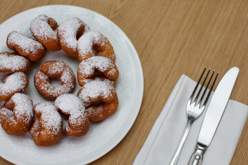 a pile of delicious sweet golden brown doughnuts (donuts) with white powdered icing sugar topping laying in a white plate on a wooden table background next to a fork and knife