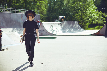 Pretty funky teenage girl carrying longboard walking in skatepark in sunglasses, hat and black clothes, for ad or blog