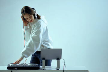 Cool young girl DJ mixes music on a mixing console and laptop, in stylish clothes, on white blue...