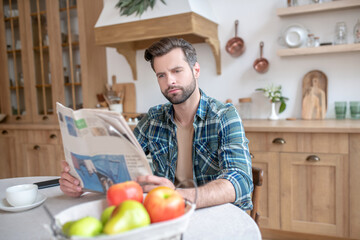 Man in a checkered shirt sitting at the table, reading newspaper and looking involved