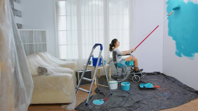 Paralyzed woman painting wall with roller brush sitting on wheelchair. Handicapped, disabled ill and immobilizes woman helping with apartment redecoration and home construction while renovating and