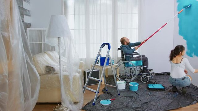 Handicapped man in wheelchair helping wife to paint apartment wall. Handicapped, disabled ill and immobilizes man helping with apartment redecoration and home construction while renovating and