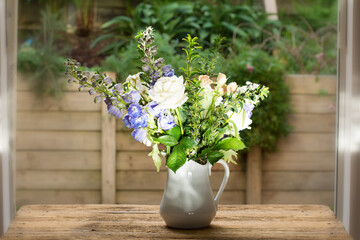 Beautiful composition with white and peach-coloured roses, blue delphoniums, and greens in a white jug on a wooden table in the garden