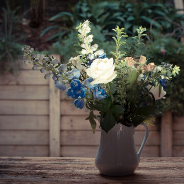 Vintage photo of a flowers arrangement with white and peach-coloured roses, blue delphoniums, and greens in a white jug against wooden background