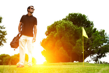 Full length portrait of stylish golfer in glasses standing on golf course with amazing flare sunset...