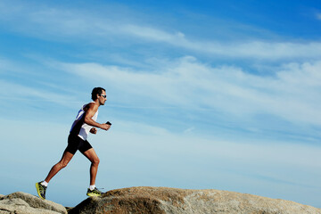 Sporty male runner jogging outdoors against copy space sky background, mature male engaged in running while listening to music in headphones, working out in mountain landscape