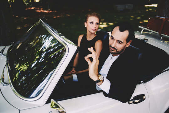 Richly dressed wealthy and famous couple sitting inside cabriolet luxury car, successful man sitting behind convertible steering wheel with its ok hand gesture, happy man and woman enjoying the life - Powered by Adobe