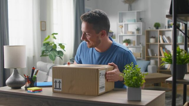 A handsome young man enters the living room with a corton box, sits at the desk and begins to open the parcel