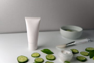 Facial Skincare white tube organic product with blank label decorate with fresh cucumber sliced and leaves next to silicone brush grey isolated background. Fresh fruit cucumber mask treatment concept