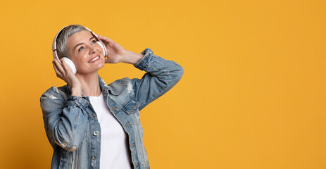 Mature woman in wireless headphones listening music over yellow background