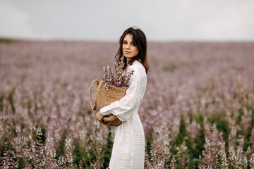 Young woman in white summer dress, standing in a big field with sage in bloom, holding a wicker bag...