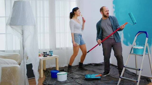 Joyful couple redecorating, dacing and singing with roller brush dipped in blue paint. Apartment redecoration and home construction while renovating and improving. Repair and decorating.
