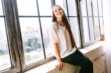 Attractive young woman in a casual wear sits on a windowsill near a huge loft-style window and laughs.