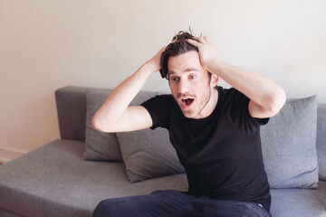 Young handsome man in black t-shirt shaking his head, watching a sport game with emotions. A guy sitting on a sofa, concept of watching TV at home