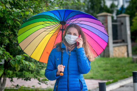 A Caucasian girl in a protective mask stands on an empty street at a bus stop under an umbrella in the spring rain and talks on the phone. Safety and social distance during a coronavirus pandemic