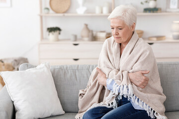 Senior lady sitting on couch covered with blanket, shivering with high temperature