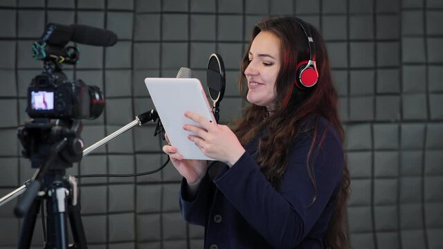 Medium shot of a female voice artist doing voice over in a sound studio. Voice actress reads text from digital tablet and dubbing to the microphone. Podcast production on the sound recording studio