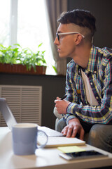 Pensive guy looks at window thoughtfully while sitting at sofa. Side view of young freelancer in working process. Man at home working remotely.