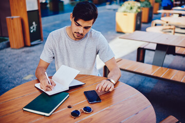 Clever male student doing homework task writing report in copybook sitting at table in cafe terrace, concentrated handsome hipster guy making plan for day noting points and schedule in dairy