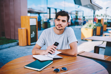 Handsome young man in casual outfit looking away while holding modern smartphone in hand and thinking on ideas for developing of own web page in internet sitting at wooden coffee table with textbooks