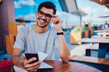 Portrait of cheerful young hipster guy in trendy eyeglasses happy receiving message with money transaction from bank, smiling handsome male student enjoying free time blogging in social networks