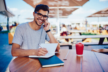 Happy bearded male student excited with getting invitation for interview noting contacts and address during phone talk, cheerful hipster guy wiring creative ideas in notepad consulting with friend