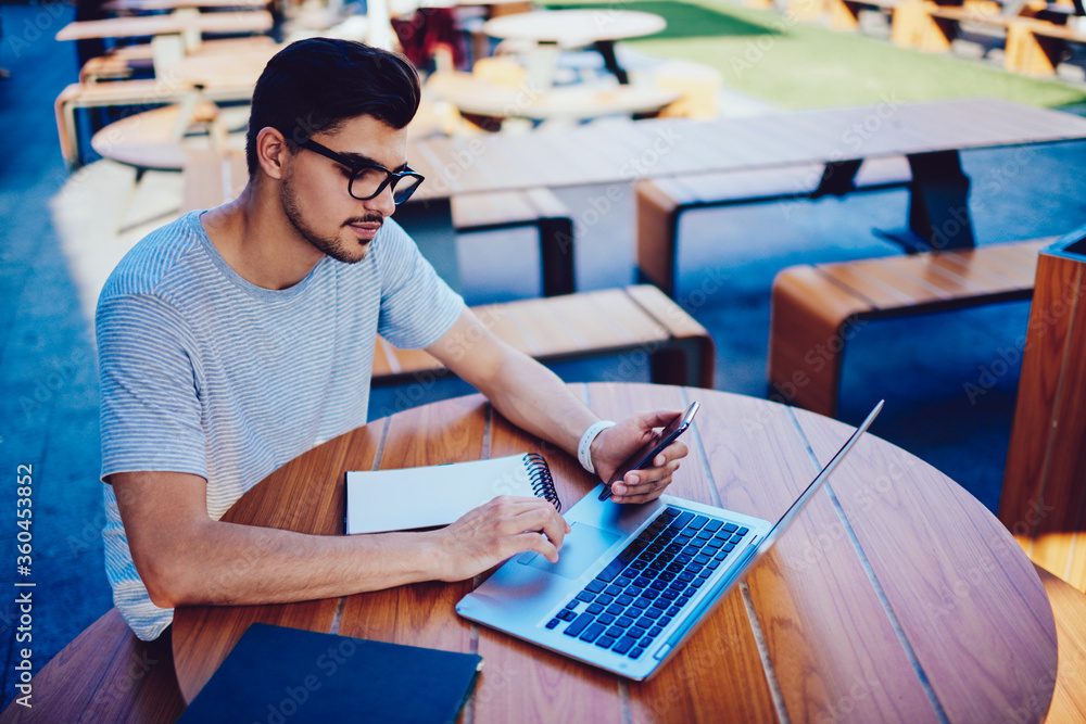 Poster concentrated male programmer in trendy eyeglasses synchronizing multimedia files from mobile with la