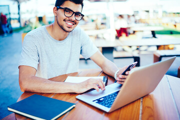 Portrait of successful male blogger in eyeglasses smiling at camera while synchronizes media files from smartphone to modern laptop computer using wireless internet connection for productive work