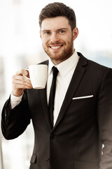 Business concept. Successful young businessman at work. Manager standing in office happy drinking coffee from cup. Man smiling in suit indoors on glass window background